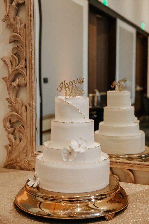 Three-tier white wedding cake with gold Happily Ever After topper and white flowers, displayed on a gold stand in front of a mirror.