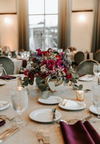 A round banquet table with a vibrant floral centerpiece, set with plates, glasses, silverware, and purple napkins.