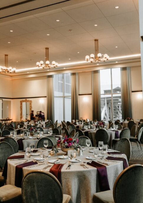 Elegant banquet hall with round tables set for an event, featuring floral centerpieces, purple napkins, and large windows letting in natural light.