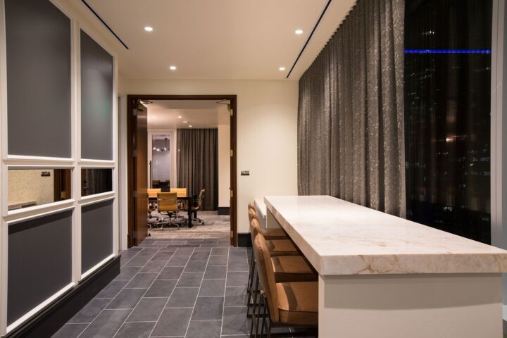 Modern office space with a marble counter and chairs, gray tile floor, large windows, and an adjoining conference room with a wooden table and chairs.