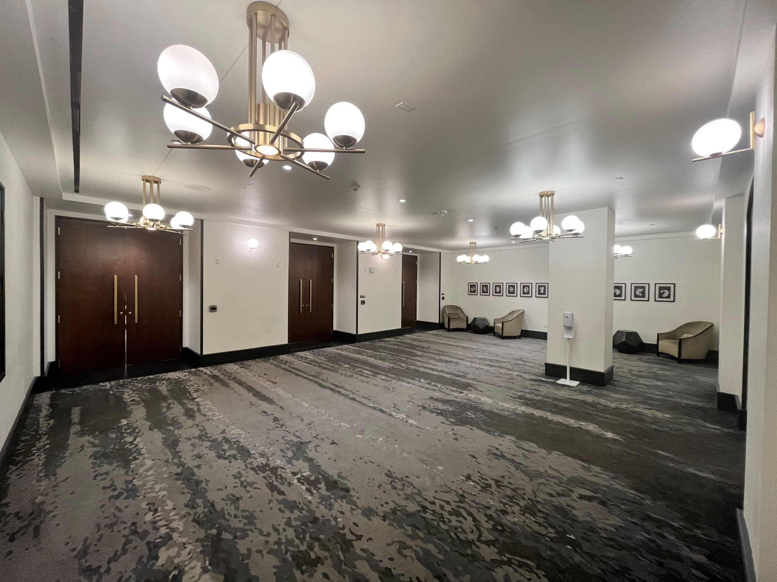 Spacious hallway with patterned carpet, modern chandeliers, wooden doors, tan chairs, framed art on the walls, and a hand sanitizer stand.