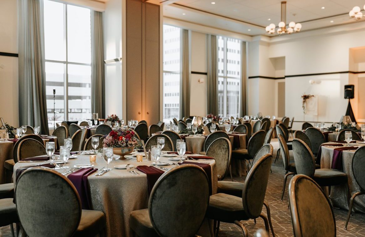 Elegant banquet hall set for an event, with round tables, gray chairs, floral centerpieces, and large windows letting in natural light.