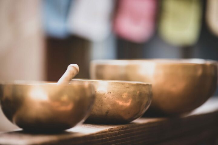 Three brass singing bowls, one with a mallet inside, rest on a wooden surface with blurred colorful backgrounds.