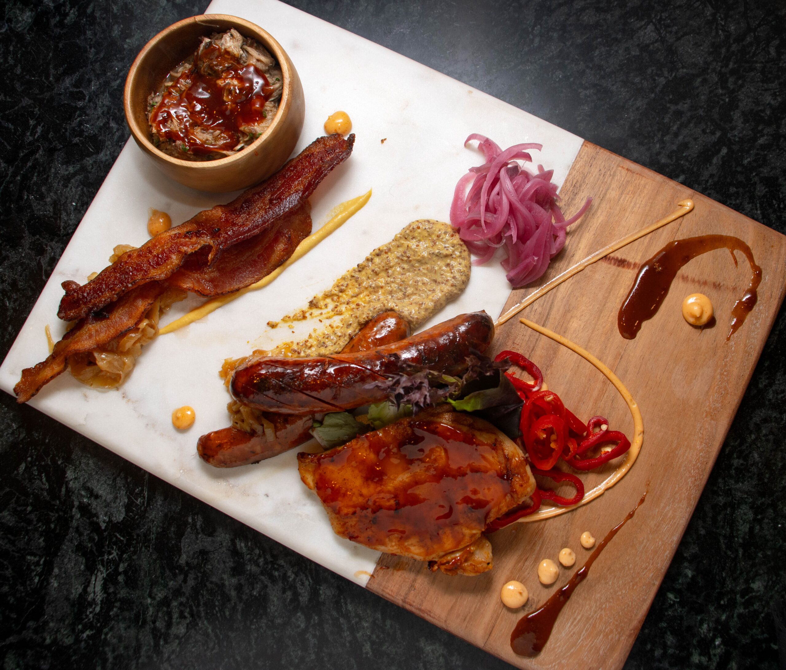 A charcuterie board with sausages, bacon, mustard, pickled onions, red peppers, sauce, and a small bowl of dip on a marble and wood platter.