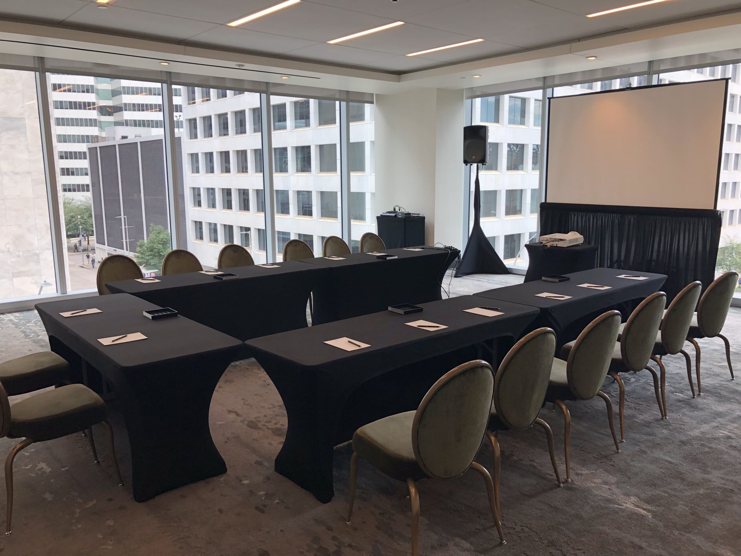 Conference room with U-shaped table setup, chairs, notepads, a projector screen, windows, and city buildings visible outside.