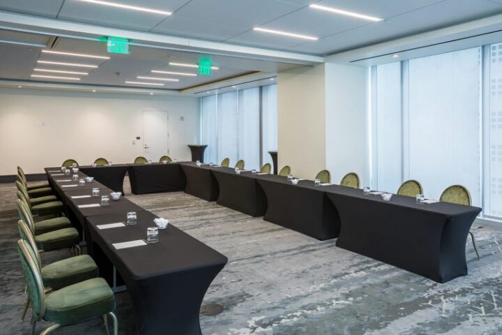 Conference room with U-shaped table setup, green chairs, water glasses, notepads, and floor-to-ceiling windows letting in natural light.