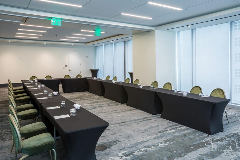 Conference room with U-shaped table setup, green chairs, water glasses, notepads, and floor-to-ceiling windows letting in natural light.