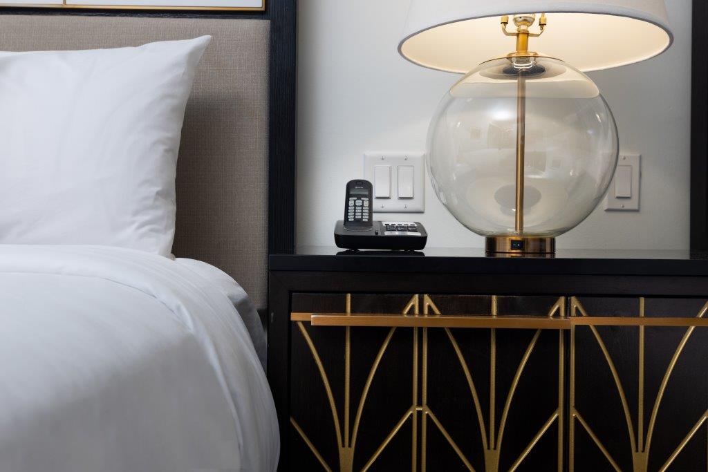 A bedside table with a glass lamp, a cordless phone, and part of a neatly made bed with white bedding.