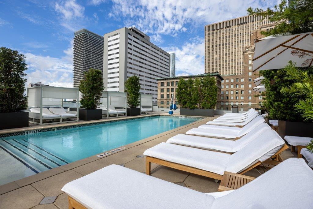 Rooftop pool with white lounge chairs, surrounded by tall buildings under a partly cloudy blue sky.