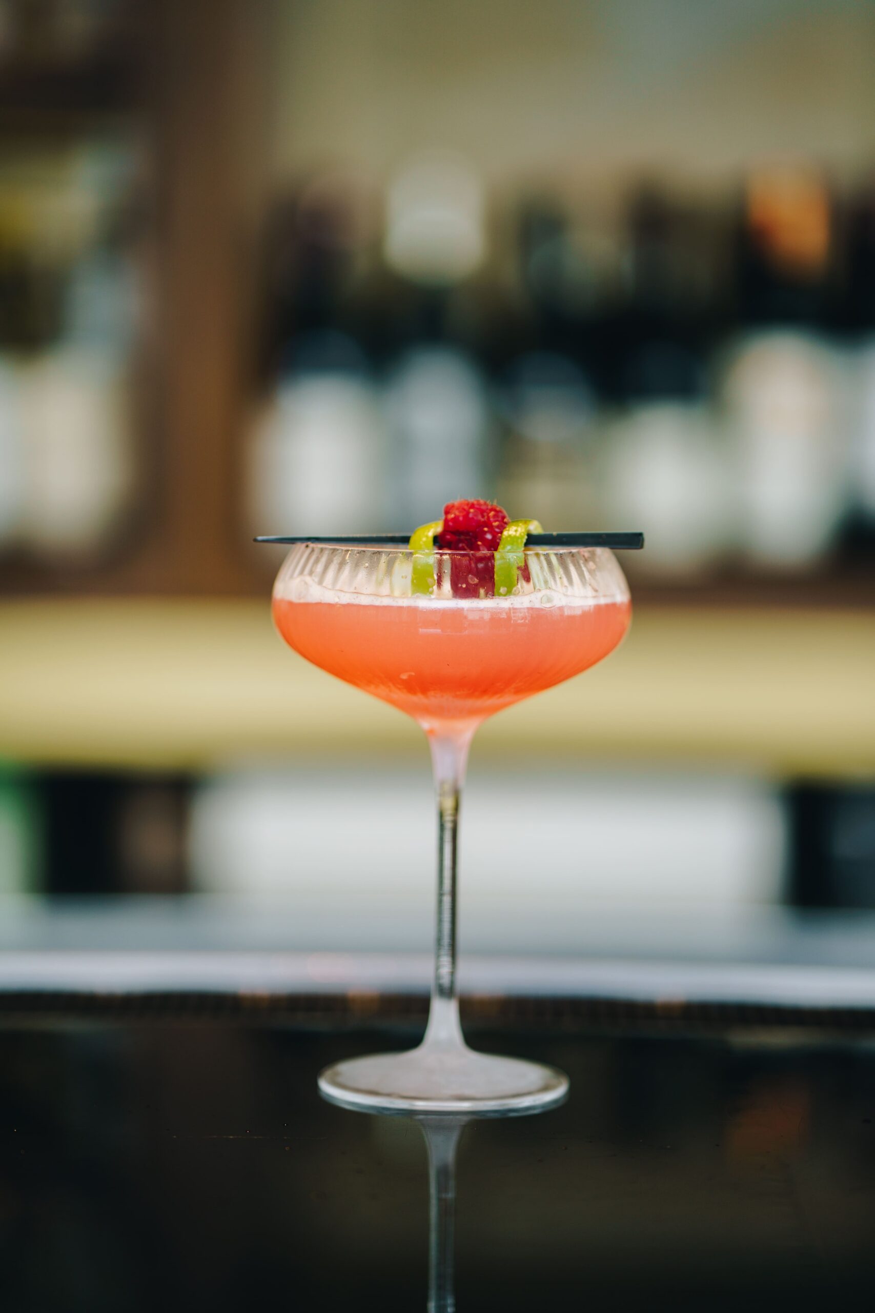 A pink cocktail in a coupe glass, garnished with a berry and a citrus twist, sits on a bar with a blurred background of bottles.
