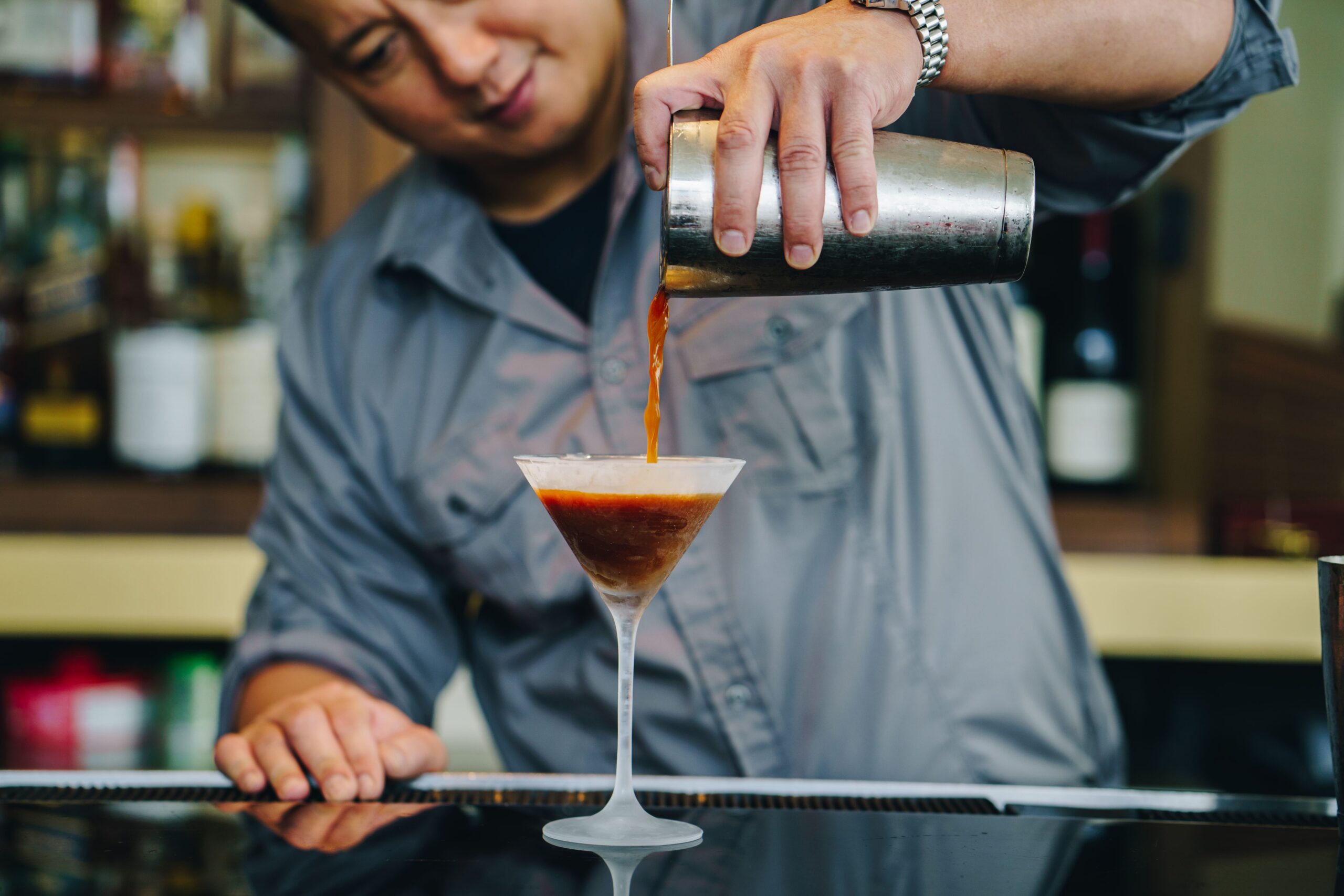 Bartender pouring a dark cocktail from a shaker into a martini glass at a bar, with bottles blurred in the background.