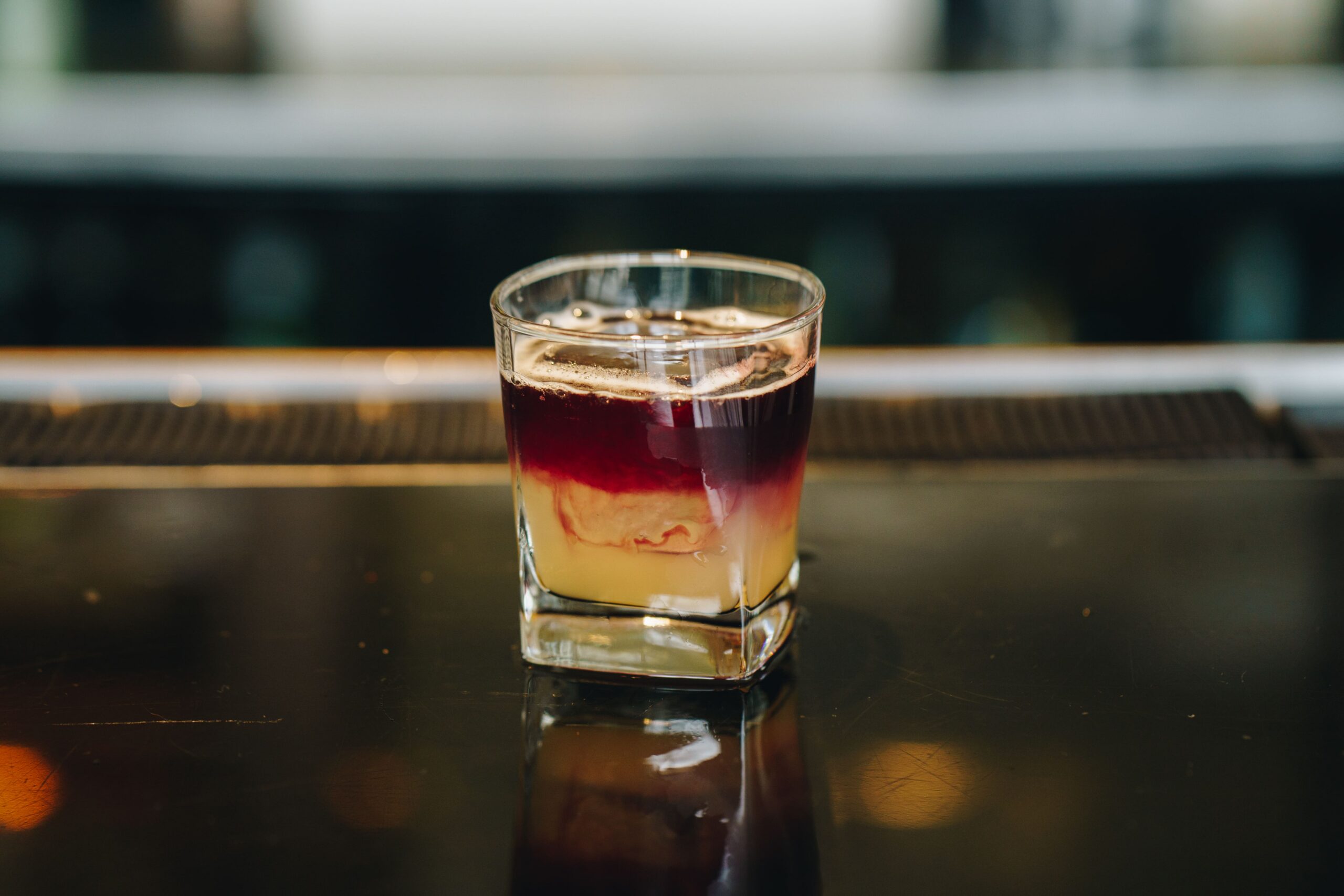 A layered cocktail in a glass, with dark red, creamy, and yellow layers, sits on a glossy bar counter.