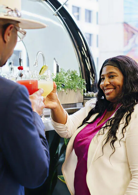 Two people clink cocktails and smile at an outdoor bar, with city buildings in the background and fresh herbs on the counter.