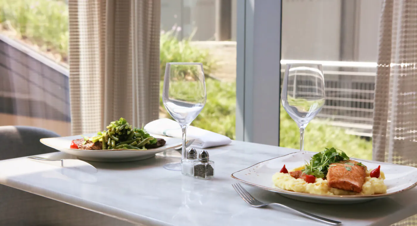 A table for two with plates of food, empty wine glasses, and utensils by a window overlooking greenery.