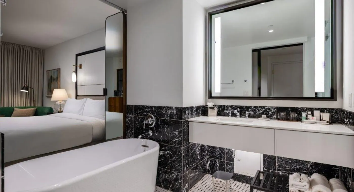 Modern hotel bathroom with a white bathtub, black marble walls, large mirror, and a view into a bedroom with a bed and lamp.