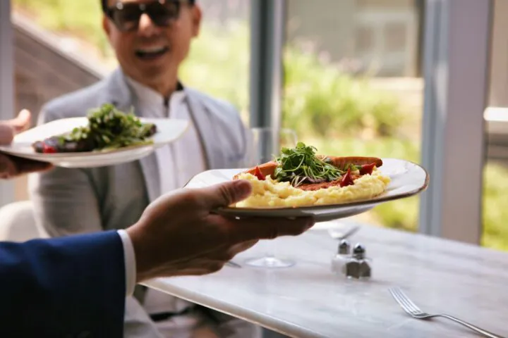 A person serves a plate of food to a man wearing sunglasses, who is smiling and sitting at a bright, sunlit restaurant table.