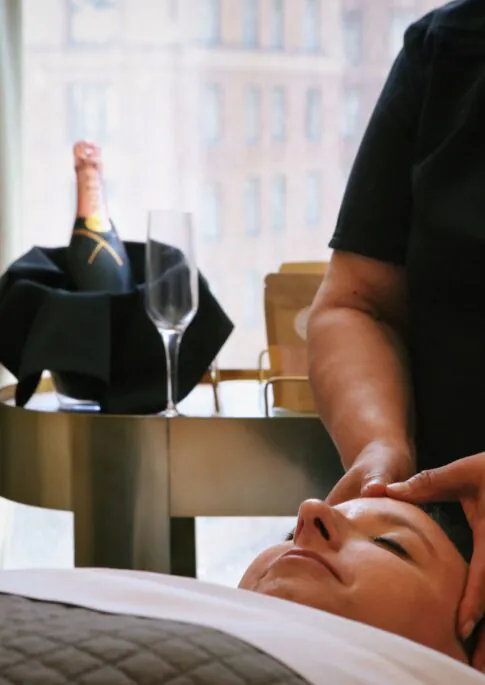 A person receives a facial massage in a spa room, with champagne and a glass on a table nearby and city buildings visible through the window.