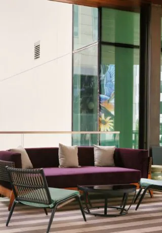 Modern outdoor lounge with a purple sofa, chairs, a small table, and a TV on the wall, with tall windows and city buildings visible outside.