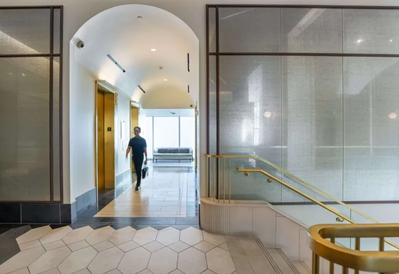 A person walks through an elegant, arched hallway with gold elevators, glass walls, and a staircase with a brass handrail in a modern office or hotel lobby.