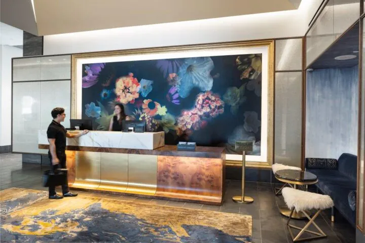 Hotel front desk with two people; a floral mural behind the counter, modern decor, gold and marble elements, and a blue seating area on the right.