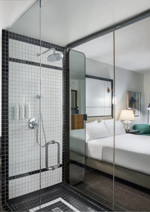 Modern hotel bathroom with a glass shower, large mirror, black and white tiles, and a view into a bedroom with a made bed and city lights outside.